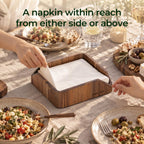 2 hands reaching for napkins from opposite sides of an acacia wood napkin holder on a dining table
